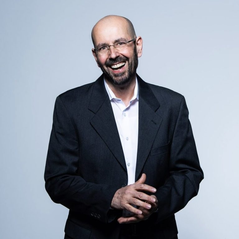 Michael Hack Lächelnder Mann in Anzug, mit kurzem Bart und Brille, stehend vor neutralem Hintergrund.